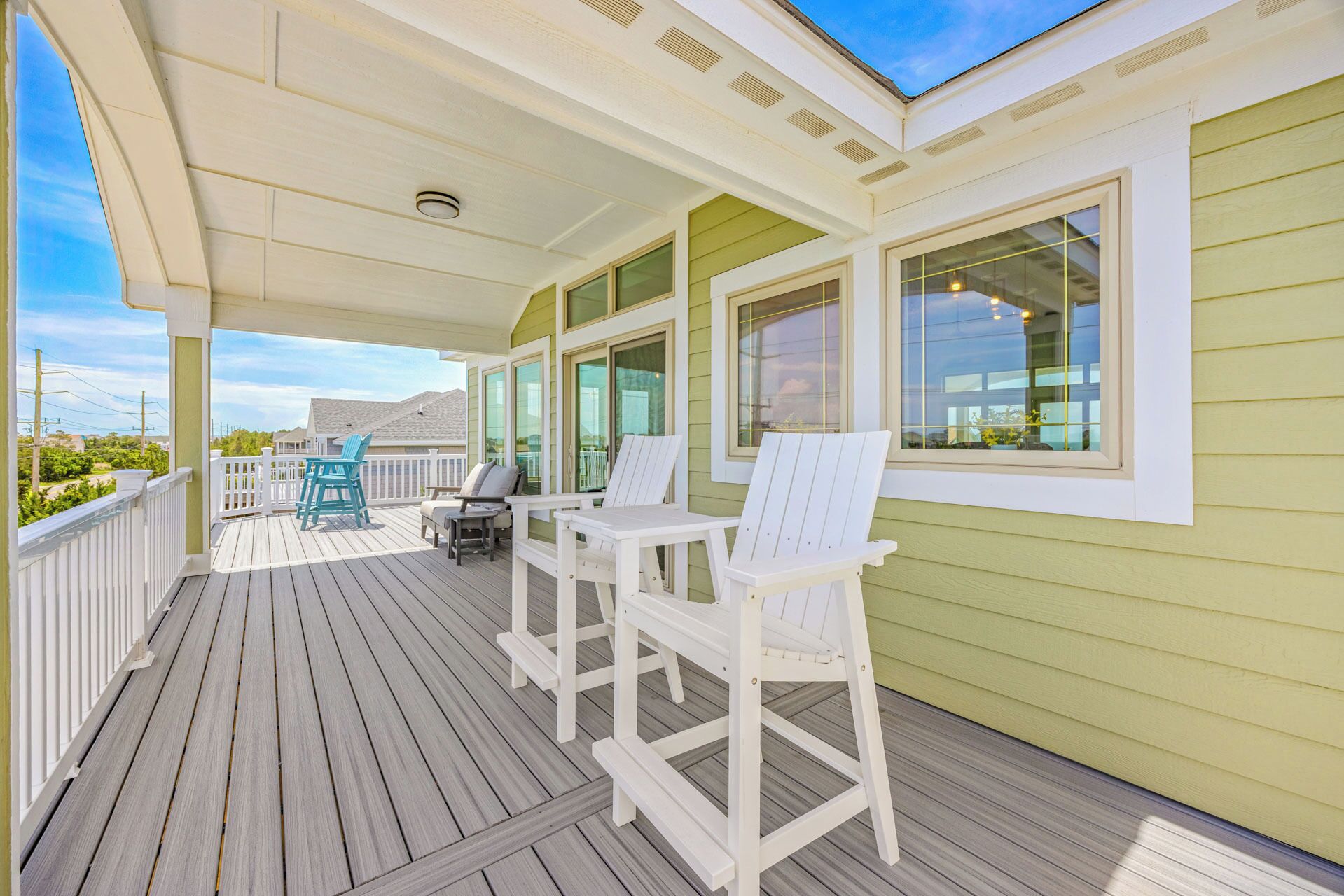 A covered outdoor deck with white Adirondack chairs and a table, large windows, and a view of the landscape.