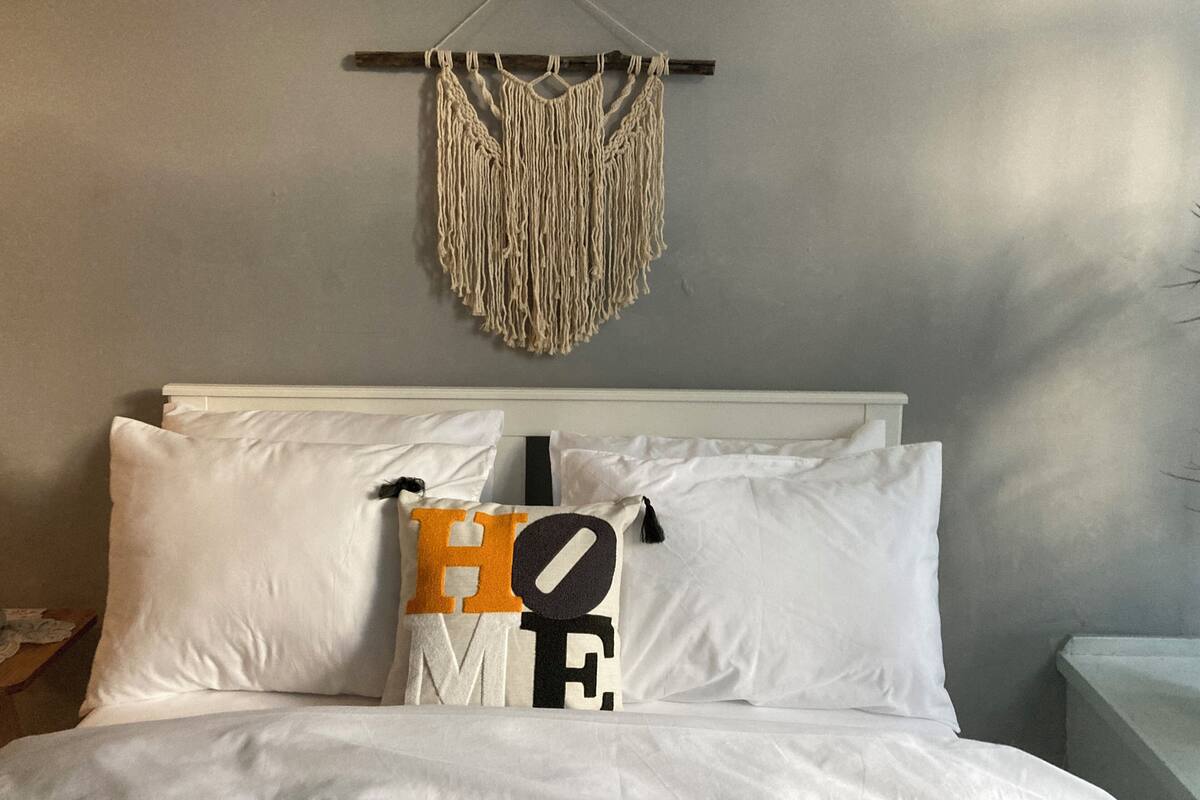 Een bed met witte beddengoed en kussens, waarvan er een het woord 'HOME' heeft, en een macramé wanddecoratie erboven.