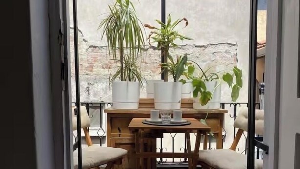 Een balkon met een houten tafel en stoelen, planten in potten en uitzicht op een bakstenen muur.