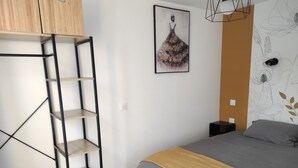 A bedroom with a bed, a wooden wardrobe, a metal shelving unit, a framed artwork, and a wall with a floral pattern.