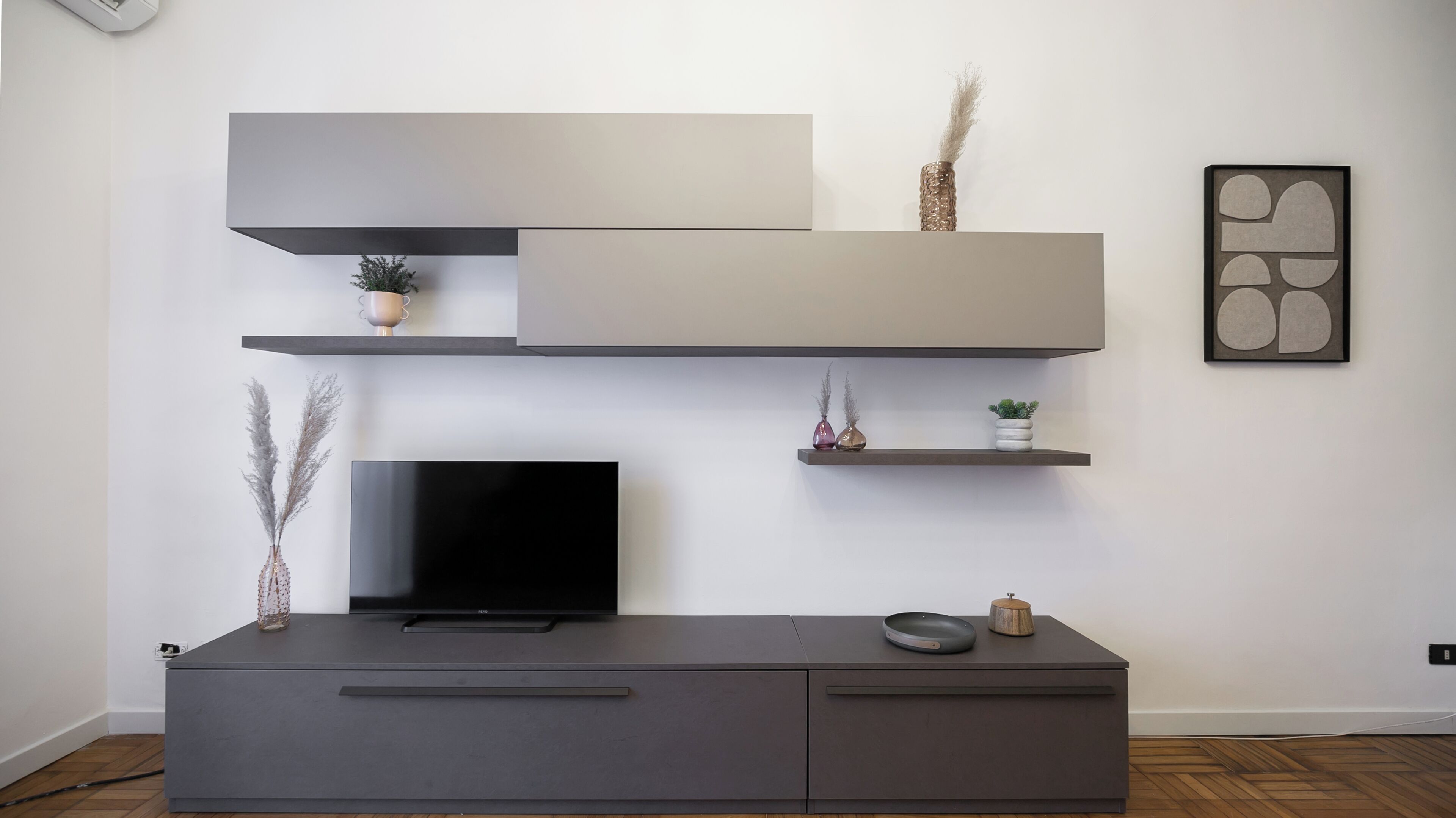 A modern living room with a flat-screen TV on a grey cabinet, two floating shelves above, and a framed abstract art piece on the wall.