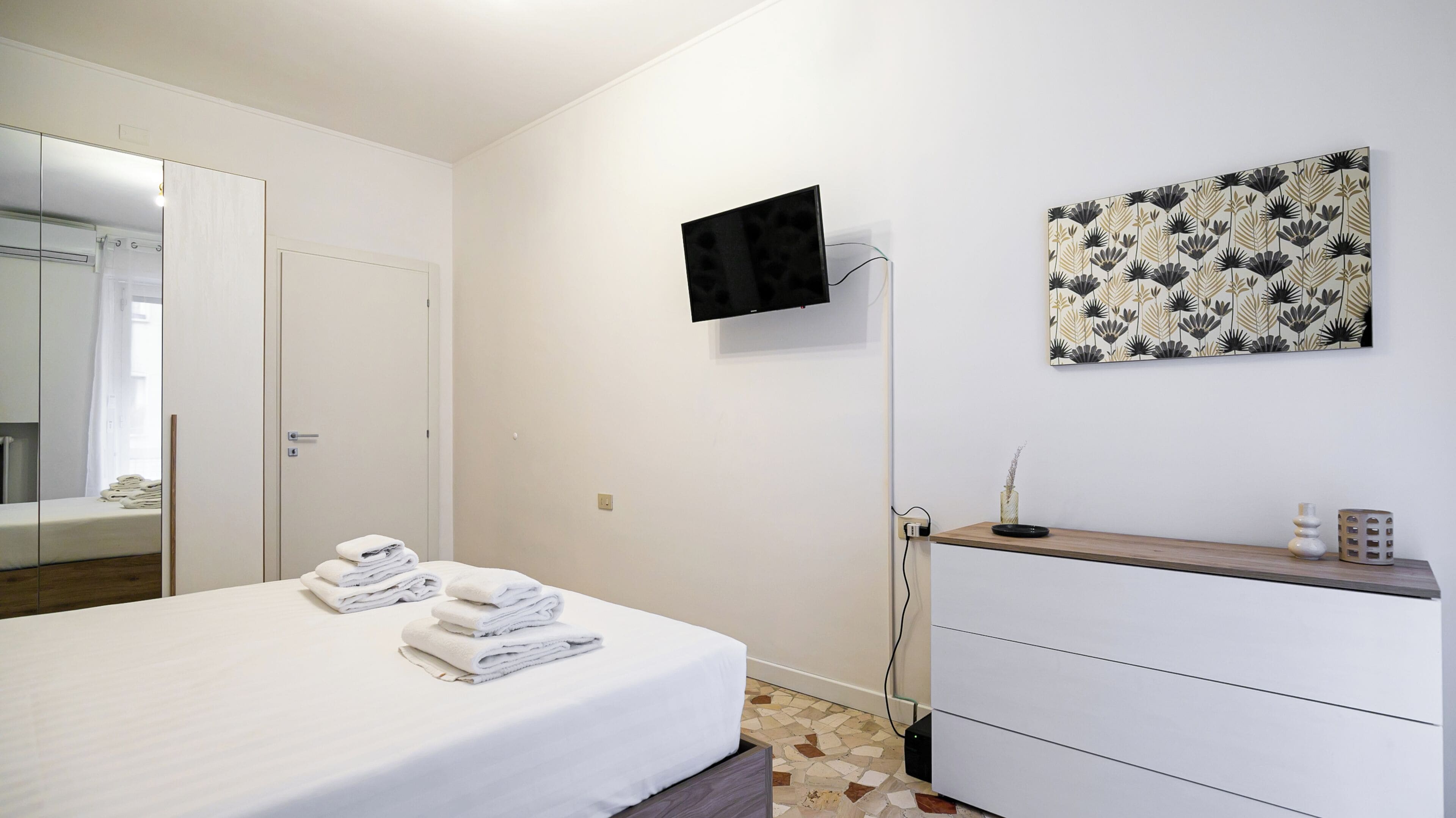 A hotel room with a bed, a bedside table with a vase, a flat-screen TV mounted on the wall, and a decorative wall piece.