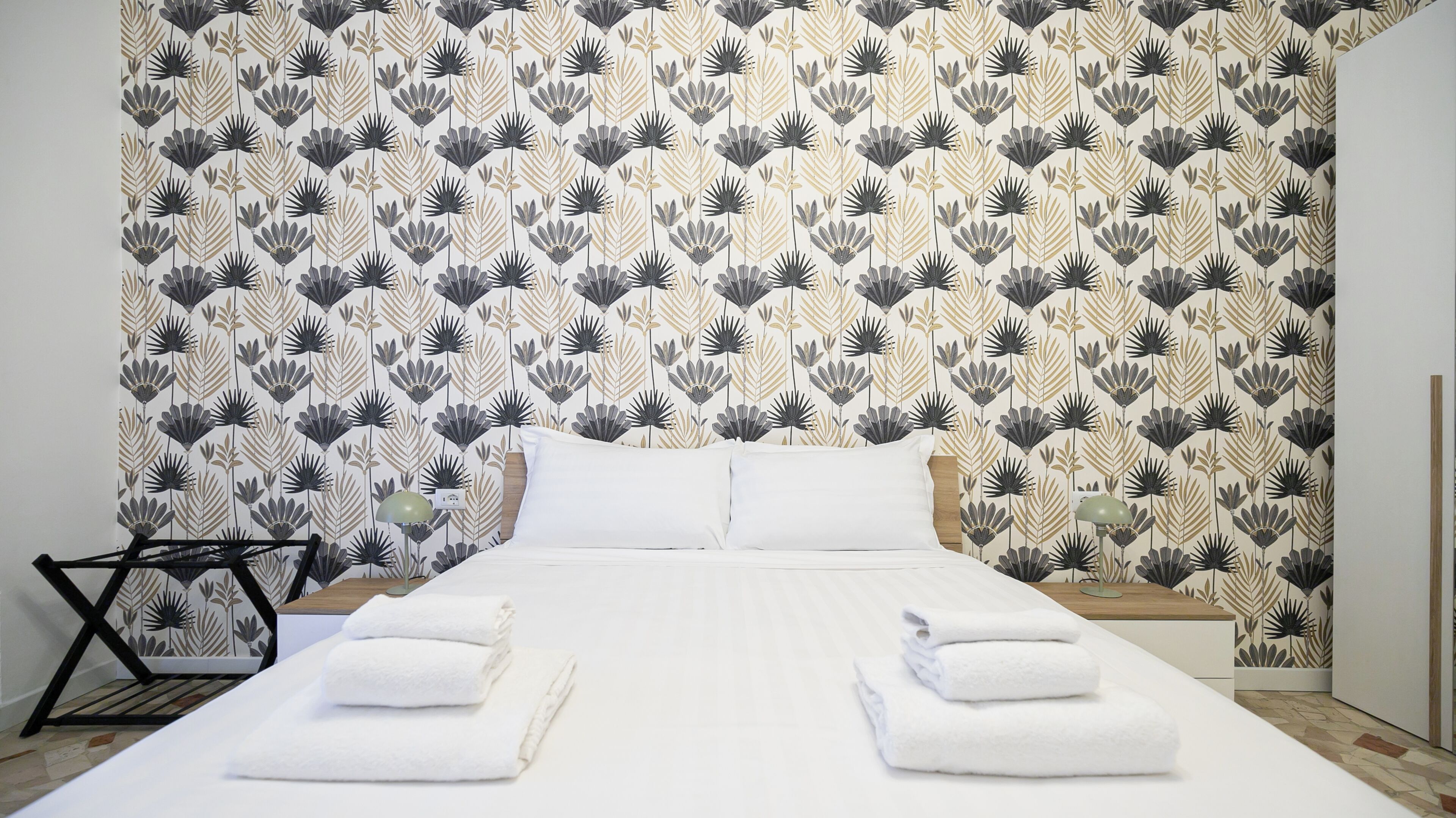 Un lit bien fait, avec du linge de lit blanc et deux serviettes pliées, une table de chevet avec une lampe, et un mur orné d’un motif floral.