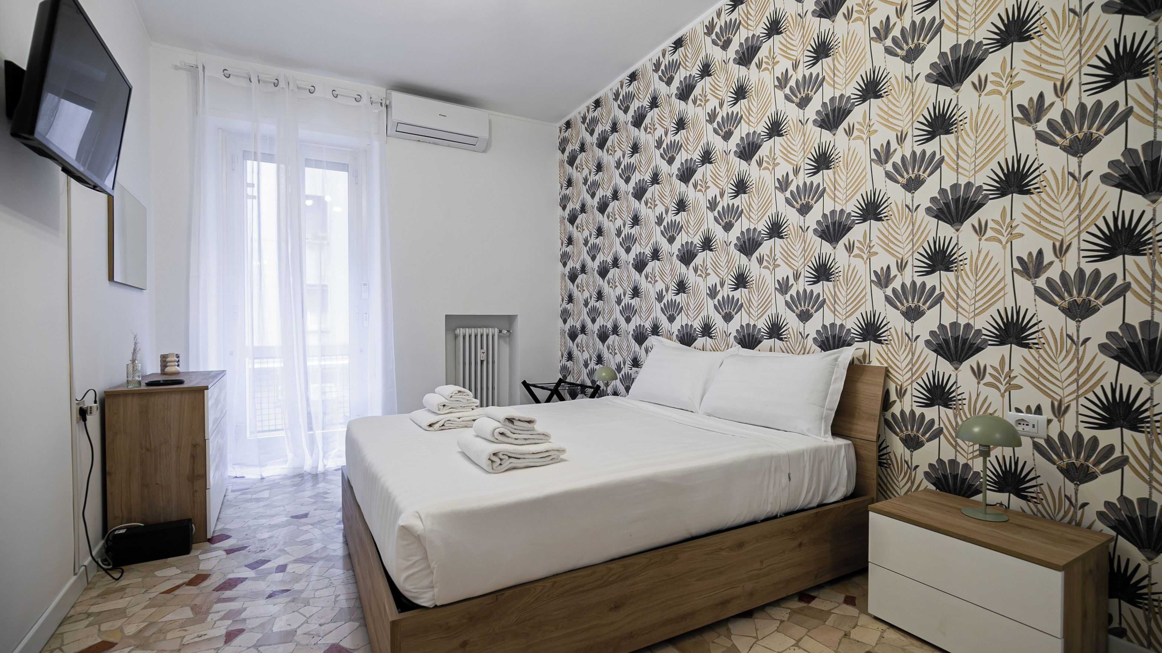 A neatly made bed with folded towels, a bedside table with a lamp, and a wall with a floral pattern.