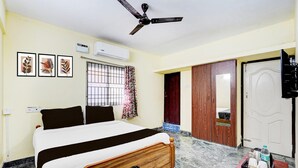 A hotel room with a bed, ceiling fan, air conditioning unit, window with curtains, wooden wardrobe, and a small table with items. - Super Collection O Sathyabama University (Chennai)