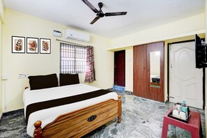A hotel room with a bed, ceiling fan, air conditioning unit, window with curtains, wooden wardrobe, and a small table with items. - Super Collection O Sathyabama University (Chennai)
