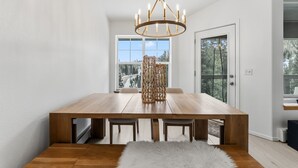 Uma área de jantar moderna com uma mesa de madeira, quatro cadeiras, um candelabro e vista para uma ponte e árvores através de grandes janelas.