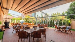Un patio al aire libre con mesas y sillas de madera, una zona de estar cubierta y vistas de los edificios y la vegetación circundantes.