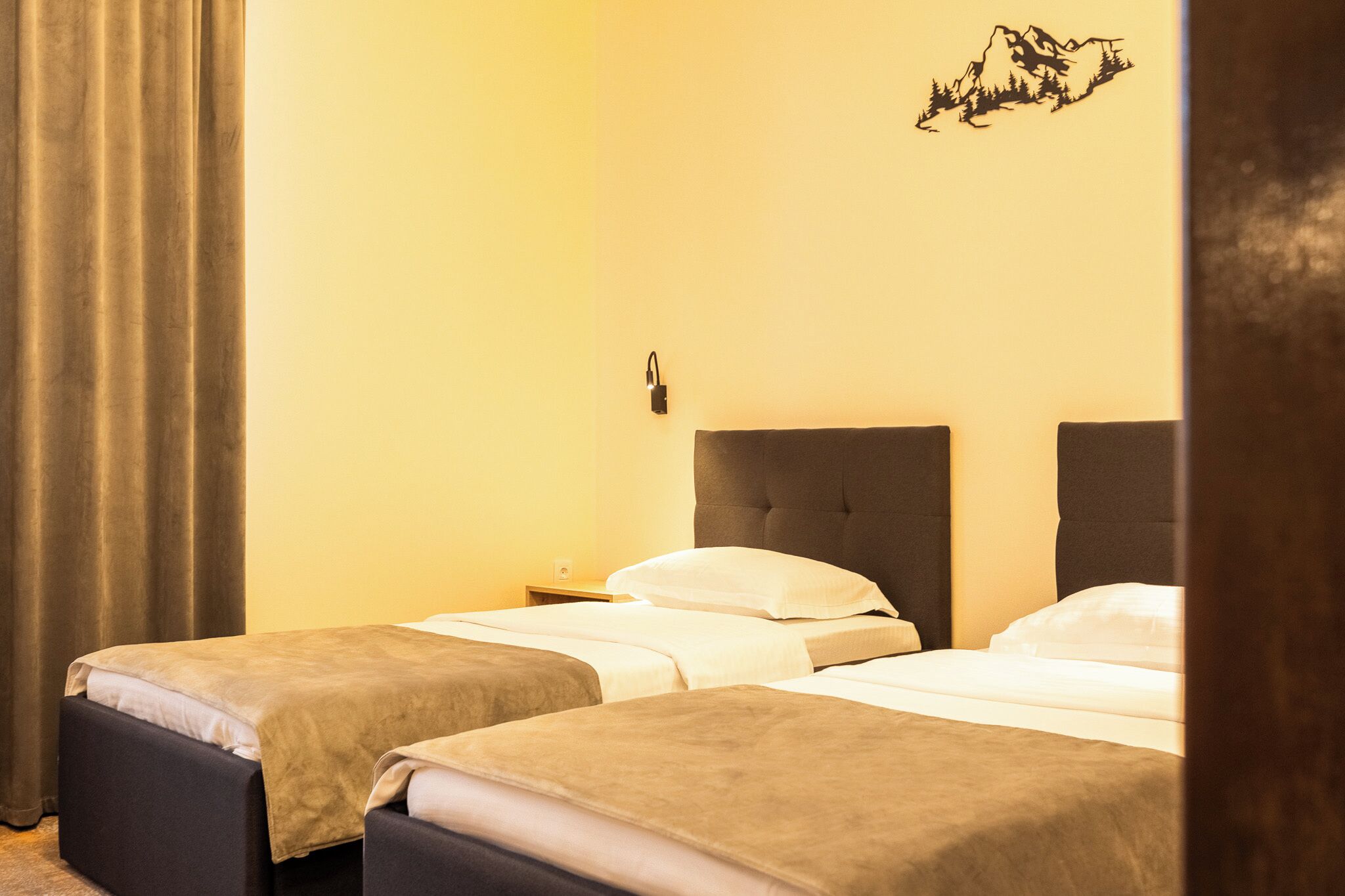 A hotel room with two single beds, a nightstand, a wall-mounted lamp, and a mountain silhouette decoration.