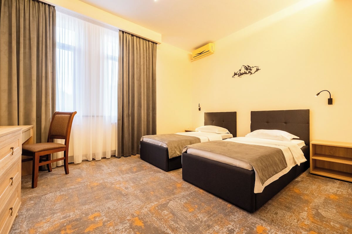 Ett hotellrum med två sängar, ett skrivbord, en stol och ett stort fönster med gardiner.