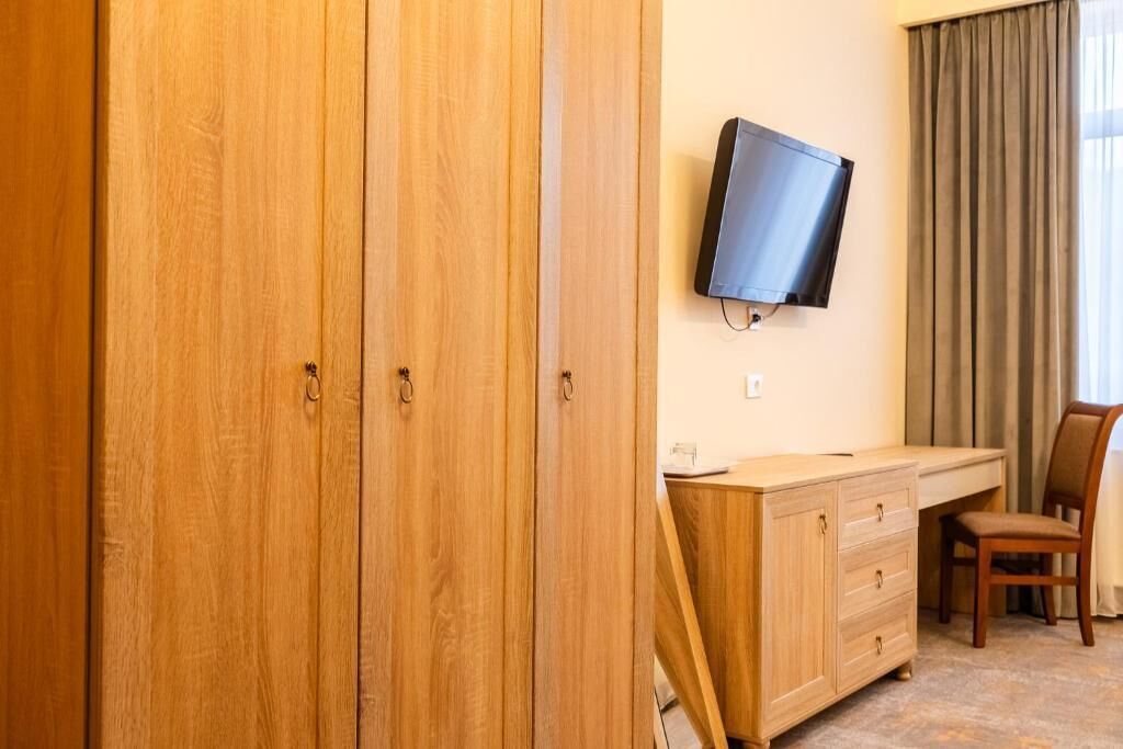 A hotel room with wooden wardrobe, a desk with a chair, a television mounted on the wall, and a window with curtains.