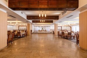 Amplia sala de comedor de hotel con múltiples mesas y sillas, paneles de madera en el techo y un escenario central con un arreglo floral.