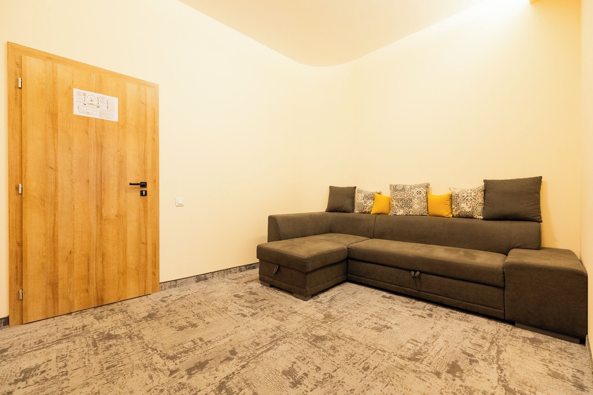 A room with a wooden door, a sectional sofa with patterned cushions, and a textured carpet.