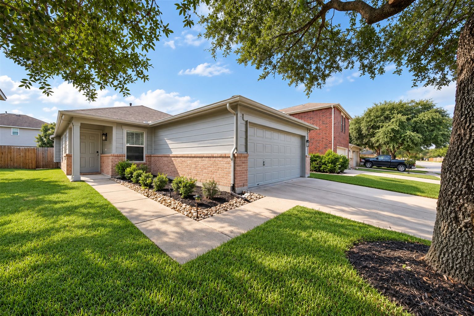 A two-story house with a garage, a front yard with a lawn and small shrubs, and a tree providing shade.