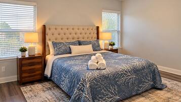 A bedroom with a tufted headboard, a blue patterned bedspread, two bedside lamps, and a pair of rolled towels on the bed.