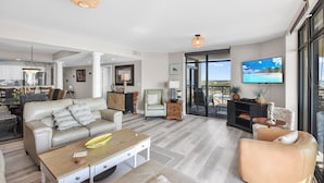 Een ruime woonkamer met een bank, salontafel en fauteuils. Een televisie is aan de muur bevestigd, en er is een balkon met uitzicht.