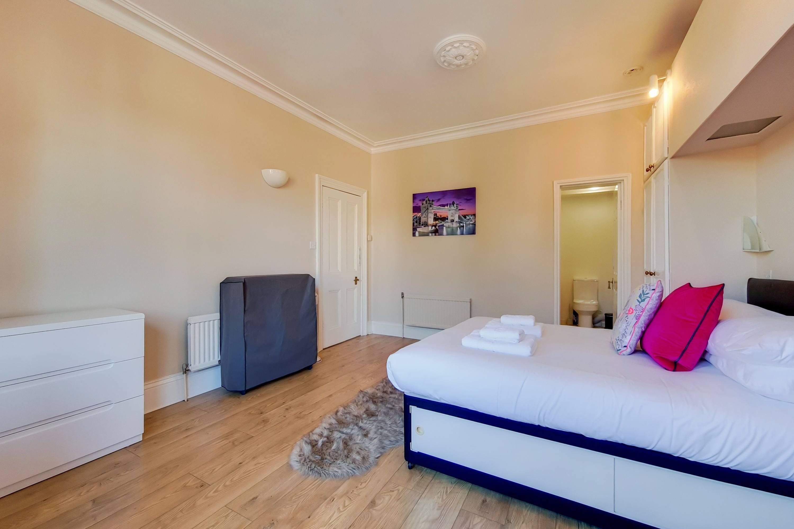 Een slaapkamer met een bed, een dressoir, een radiator, een grijs kastje en een schilderij aan de muur.