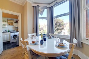 A dining area with a round table, chairs, and plates of food. A washing machine and shelves with items are visible in the background. - Apartment w Workspace in Ealing (London)