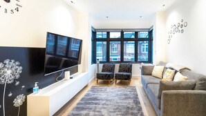 A modern living room with a flat-screen TV, two patterned chairs, a grey sofa, and a large window with a view of buildings. - Modern Duplex in Ealing Common (London)