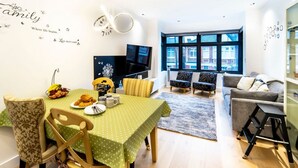 A modern living room with a dining area, featuring a green tablecloth, wooden chairs, a flat-screen TV, and a large window offering a view of buildings. - Modern Duplex in Ealing Common (London)