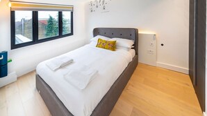 A neatly made bed with white linens and a single decorative pillow, a window with a view of buildings and trees, and a wooden floor. - Modern Duplex in Ealing Common (London)
