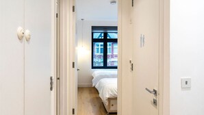 A narrow hallway with a wooden floor leading to a bedroom with a bed and a window. - Modern Duplex in Ealing Common (London)