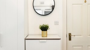 Eine weiße Tür mit Messinggriff, ein weißes Schränkchen mit einer goldverzierten Vase, in der sich eine grüne Pflanze befindet, und ein runder Spiegel an der Wand.