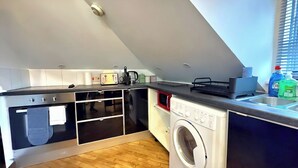 A compact kitchen with a built-in oven, microwave, and washing machine, featuring a wooden floor and a window with blinds. - Quiet Top-floor Retreat (London)