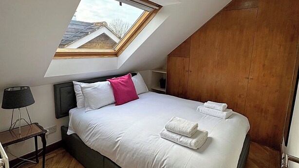 A bedroom with a skylight, a bed with white linens, a wooden headboard, a bedside table with a lamp, and a wooden wardrobe. - Quiet Top-floor Retreat (London)