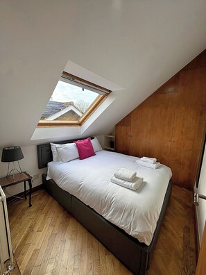 A bedroom with a skylight, a bed with white linens, a wooden headboard, a bedside table with a lamp, and a wooden wardrobe. - Quiet Top-floor Retreat (London)