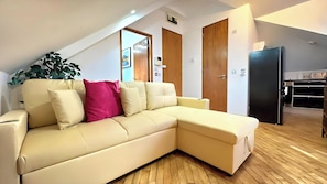 Un salon moderne avec un canapé d’angle beige, un plancher en bois et un coin cuisine doté d’un réfrigérateur noir.