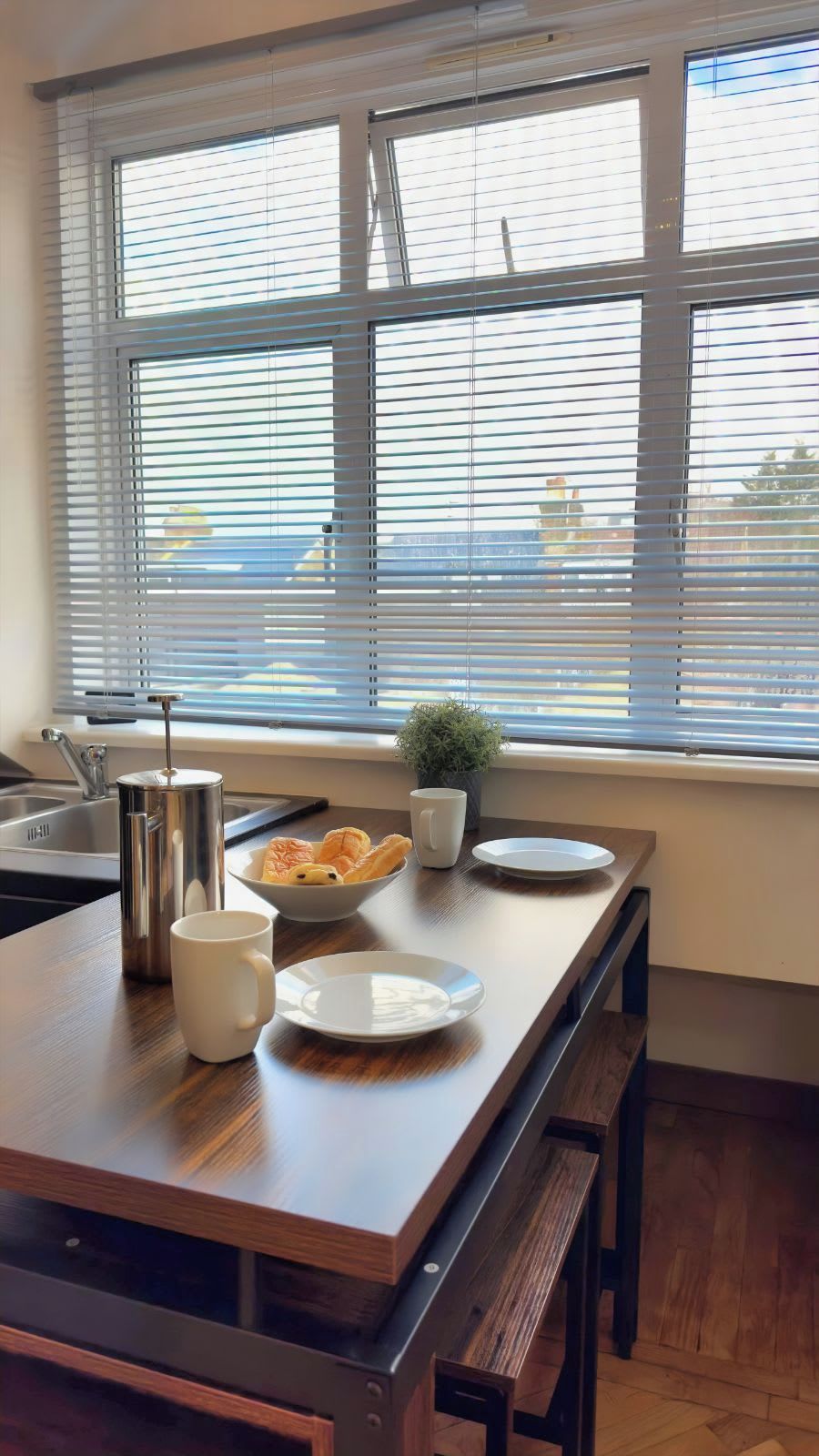 A kitchen with a wooden island, a sink, a kettle, a mug, a bowl of pastries, and a small plant.
