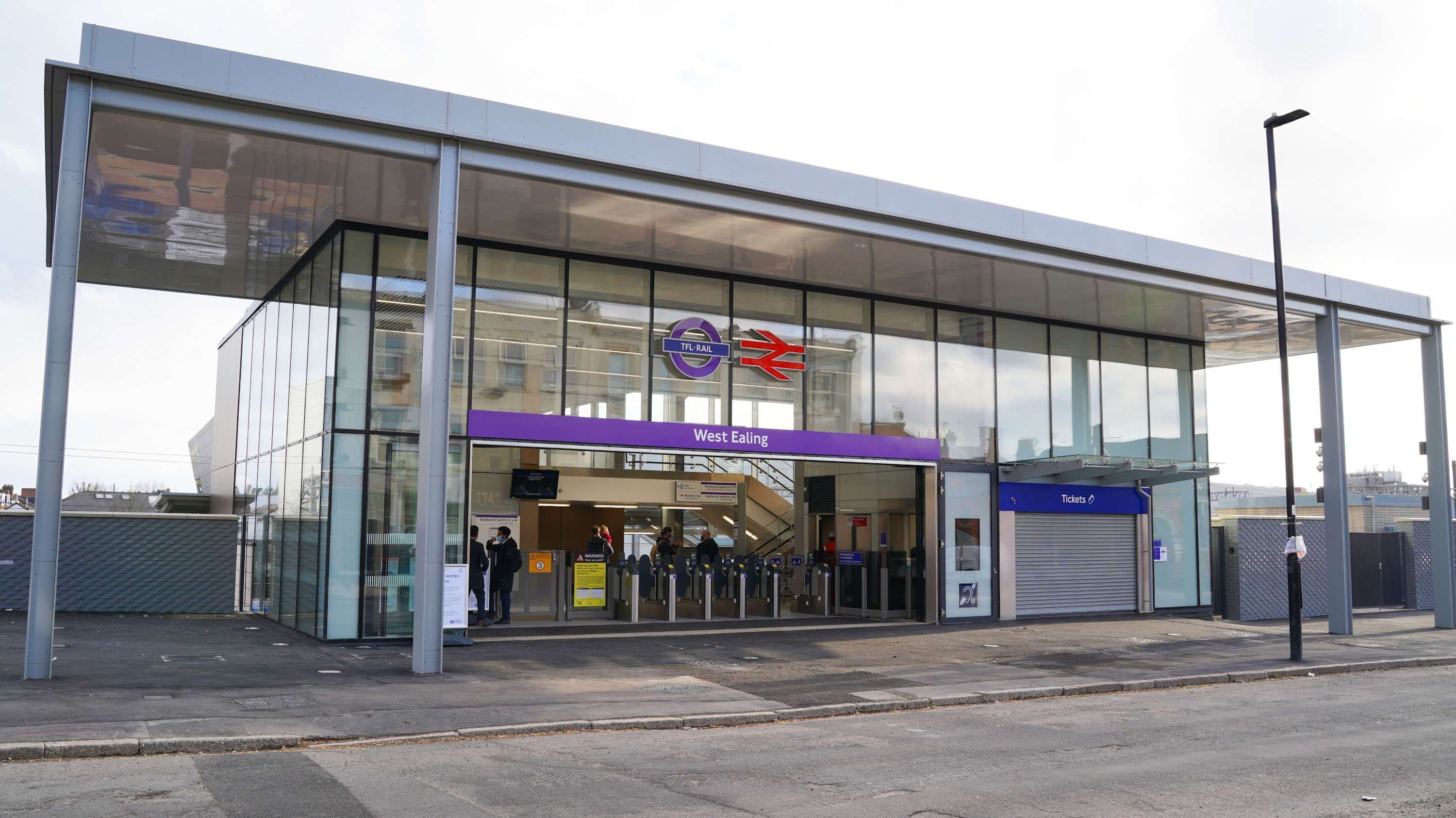 Een modern treinstation met een glazen gevel, een bord met 'West Ealing' en een loket voor 'Tickets'.