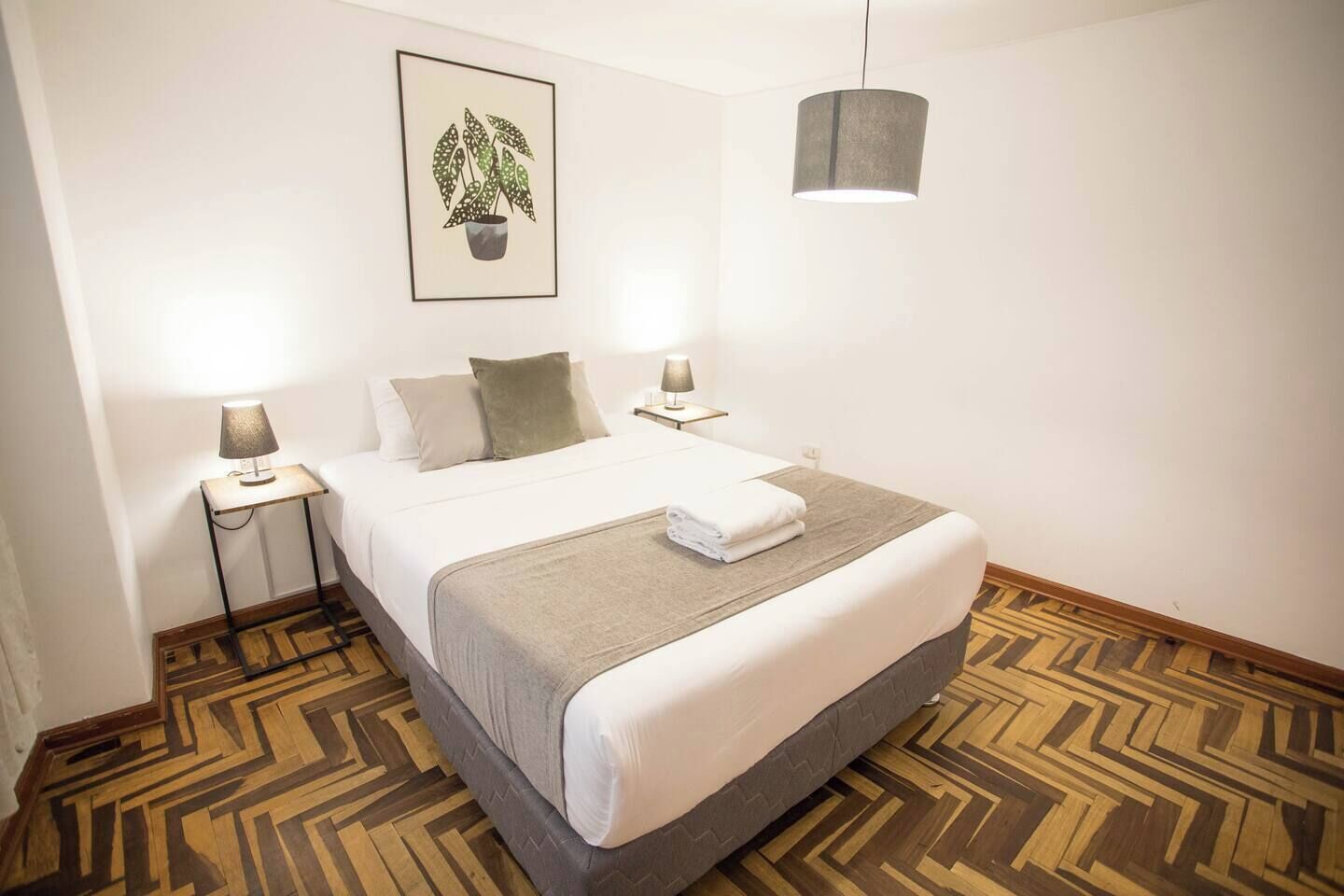 A neatly arranged hotel room with a large bed, two bedside tables with lamps, a hanging light fixture, a framed plant on the wall, and a patterned wooden floor.