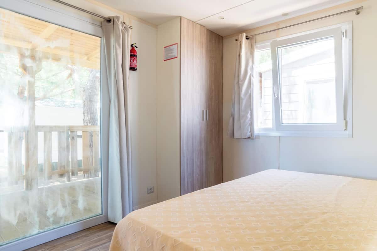 A room with a bed, a window with curtains, a fire extinguisher, and a door leading to an outdoor area.