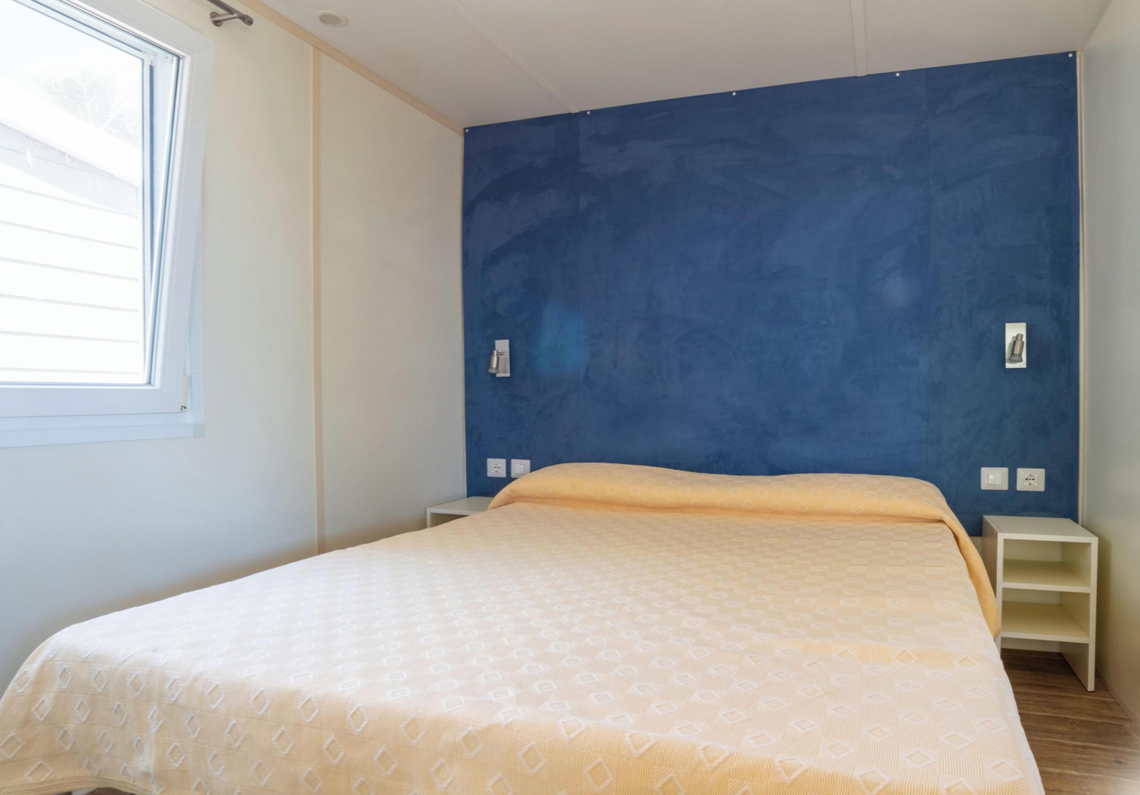 A bedroom with a bed, a window, a blue wall, and a white nightstand.