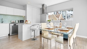 Una cocina moderna con armarios blancos, lavavajillas de acero inoxidable y una mesa de comedor de vidrio con sillas.