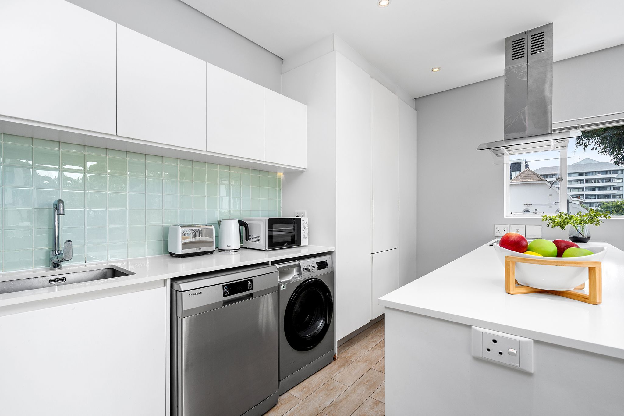 Een moderne keuken met witte kasten, een roestvrijstalen vaatwasser, een ingebouwde oven en een magnetron. Er is een raam met uitzicht op gebouwen.