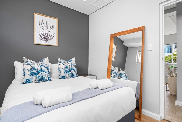 Ein modernes Schlafzimmer mit einem großen Bett, floralen Kissen, einem gerahmten Kunstwerk und einem Spiegel, der den Raum wiedergibt.