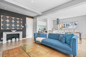 A modern living room with a blue sofa, a fireplace, and a kitchen in the background. - Vernor Court 4 (Cape Town)