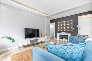 Un salon moderne doté d’un téléviseur à écran plat, d’une console blanche, d’un canapé agrémenté de coussins à motifs, d’une cheminée et d’une porte ornée d’un panneau décoratif.