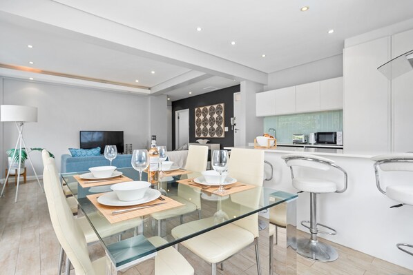 A modern living room with a glass dining table set for four, a sofa, a TV, and a kitchen area with white cabinets and a microwave. - Vernor Court 4 (Cape Town)