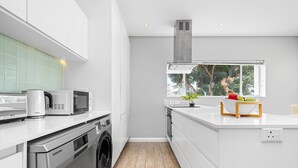 Una cocina moderna con muebles blancos, lavadora integrada, microondas y ventana con vistas a árboles.