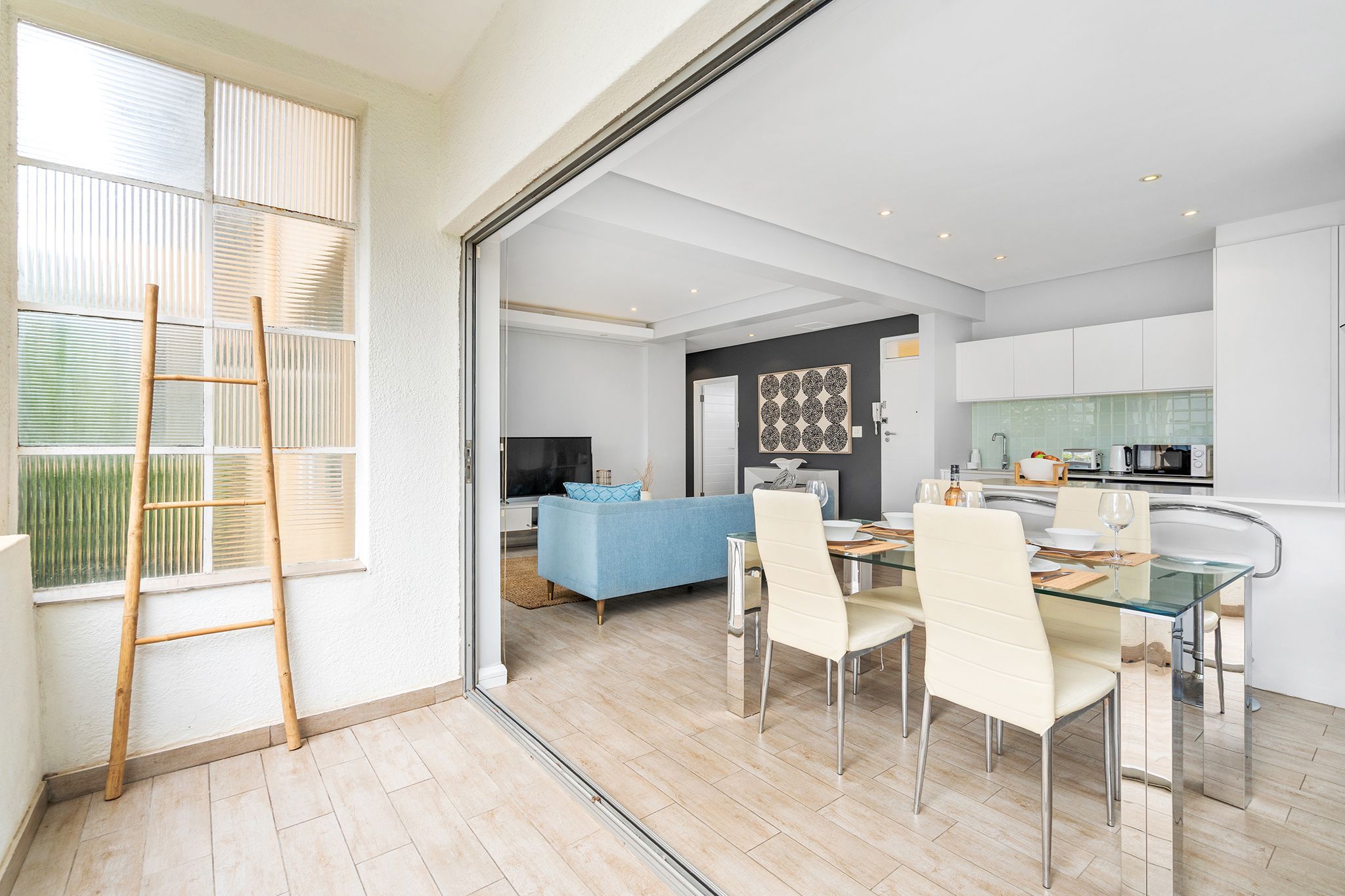 A modern kitchen with white cabinets, a dining area with a glass table, and a blue sofa in the living room.