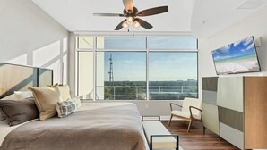 Ein modernes Schlafzimmer mit einem großen Bett, einem Deckenventilator, einem Fernseher und Blick auf die Stadt durch große Fenster.