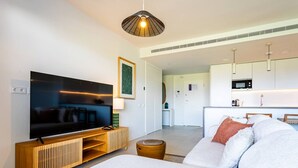A modern living room with a flat-screen TV, a wooden entertainment unit, a sofa with cushions, and a kitchen area in the background. - Ipanema 2 BR apartment with views (Mijas)