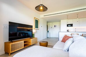 A modern living room with a flat-screen TV, a wooden entertainment unit, a sofa with cushions, and a kitchen area in the background. - Ipanema 2 BR apartment with views (Mijas)