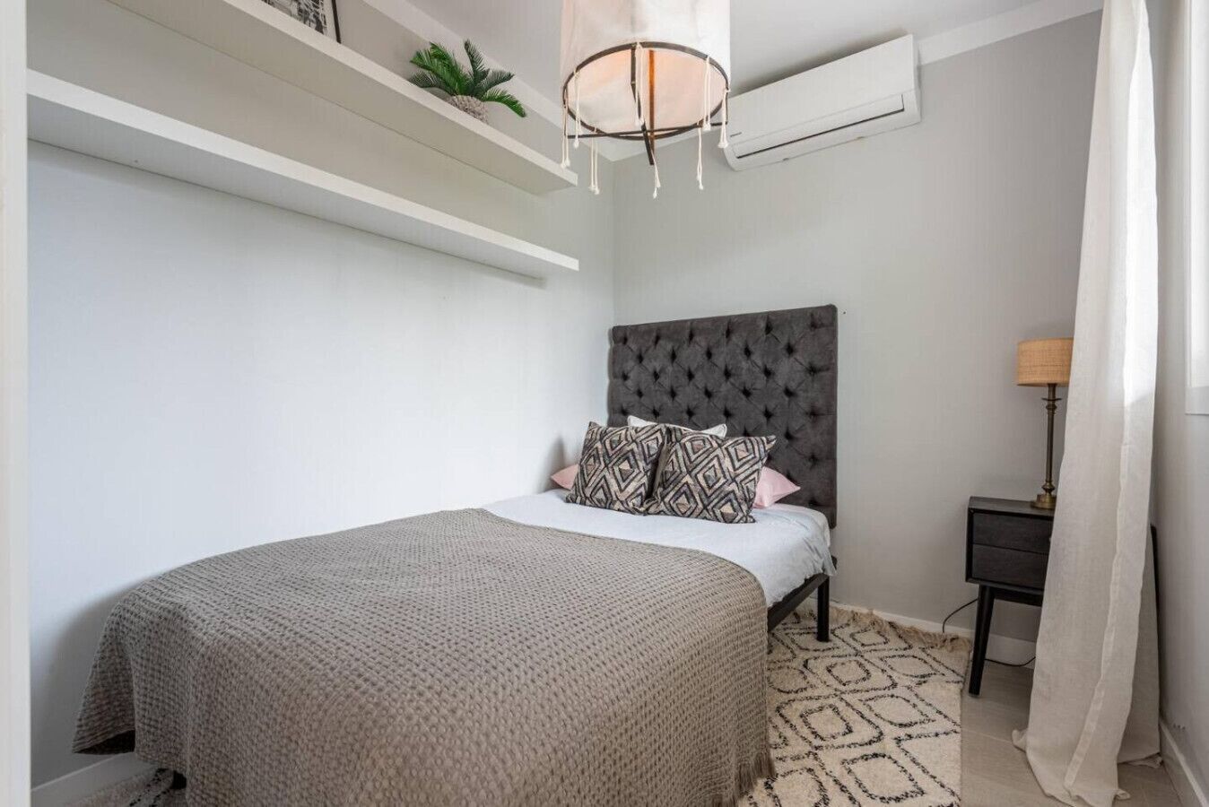 Un dormitorio con cama, cabecero, mesita de noche, lámpara y una alfombra estampada.