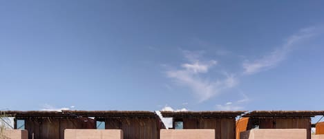 A row of rustic, wooden cabins with metal roofs, set in a dry, gravelly landscape under a clear blue sky.
