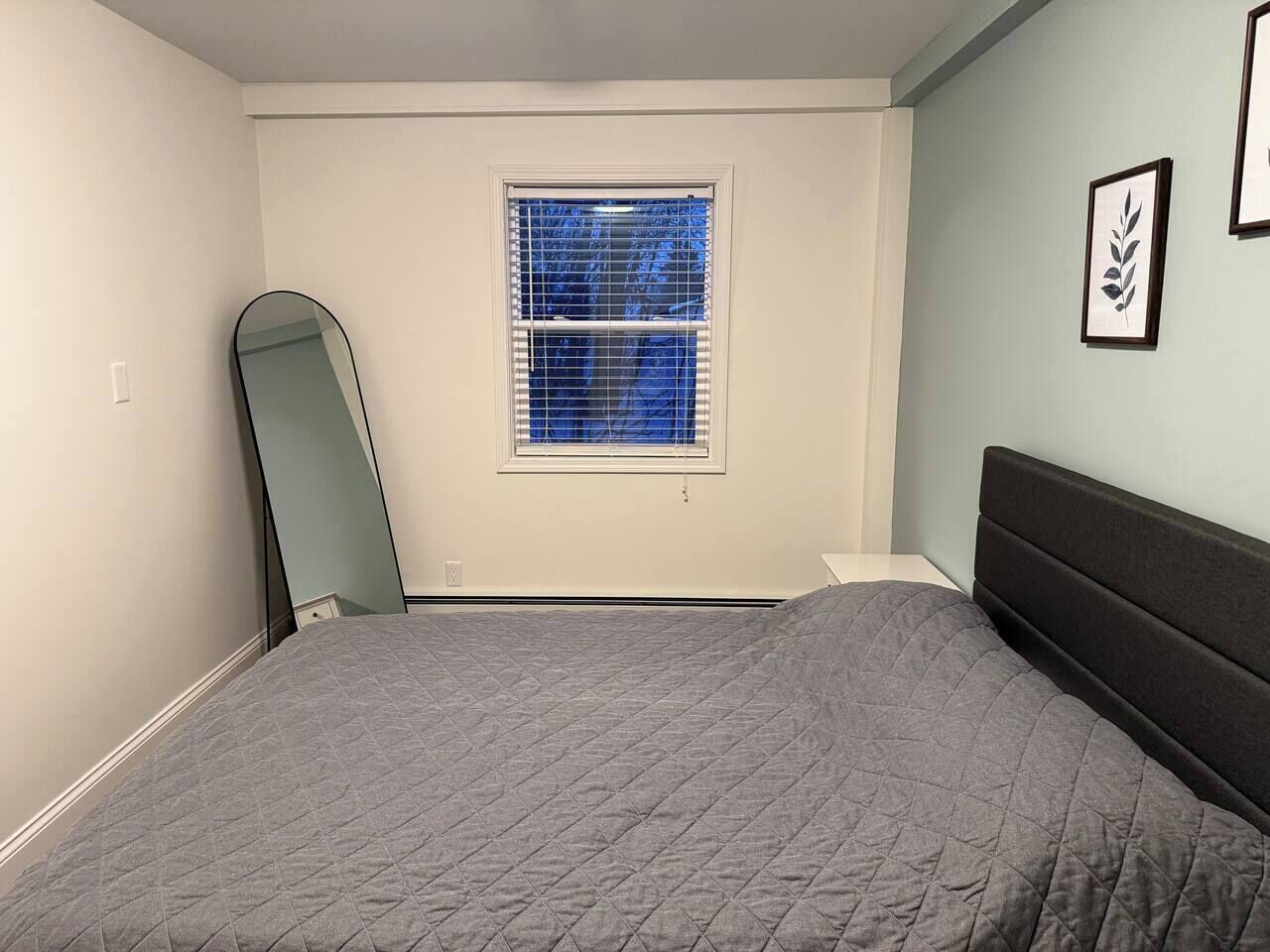 Ein Schlafzimmer mit einem Bett, einem Fenster mit Jalousien, einem Spiegel und gerahmten Kunstwerken an der Wand.